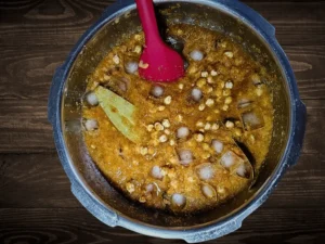 Put the puree of herbs made in the previous step, Bay Leaf, Black Cardamom, Asafoetida (Hing), Chhole Masala, Coriander Powder, Turmeric Powder, Roasted Cumin Powder, Cinnamon Powder, Water, Ice cubes and Cloves into the pressure cooker in which we had already added chickpeas.
